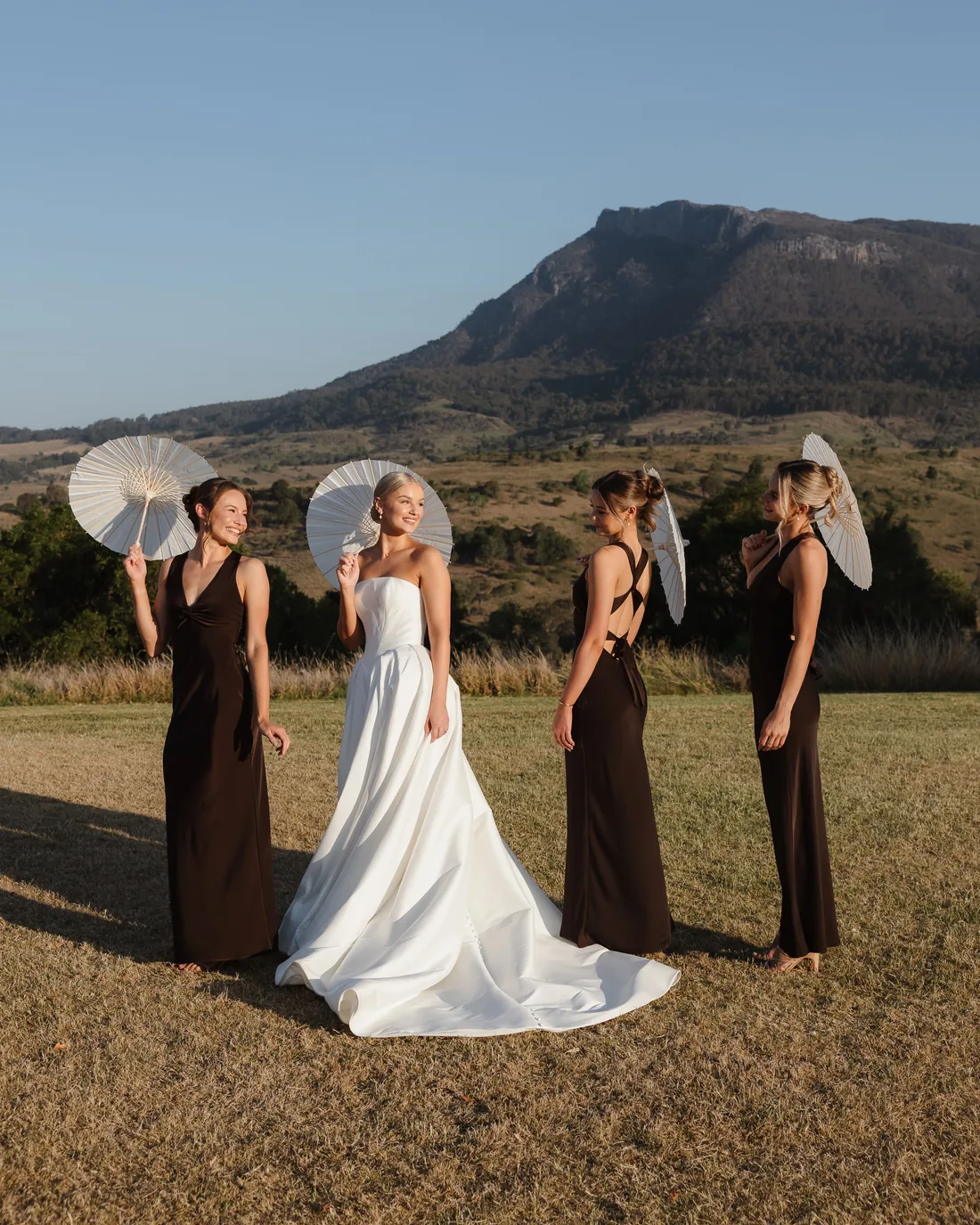 Bride and bridesmaids holding white parasols in a golden paddock, mountain backdrop
