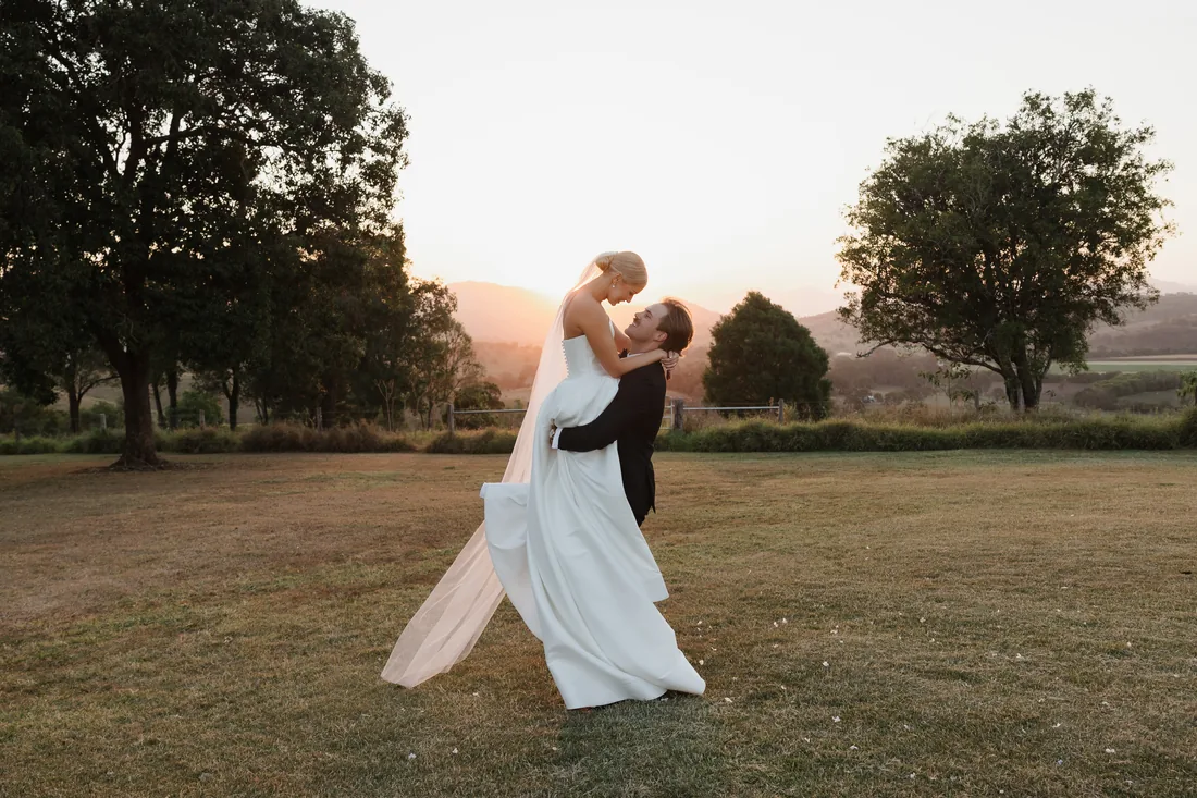 Groom lifting his bride at sunset, veil trailing, mountain silhouette behind