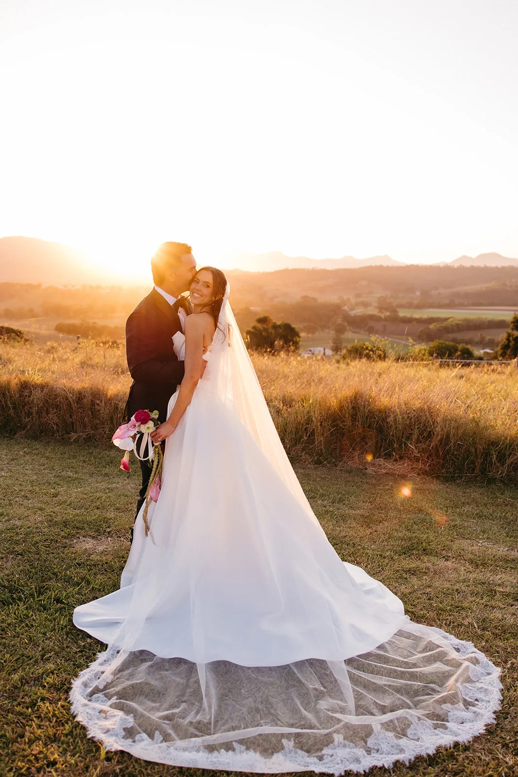 Ally and Braydon kissing at golden hour in a paddock, veil catching the light