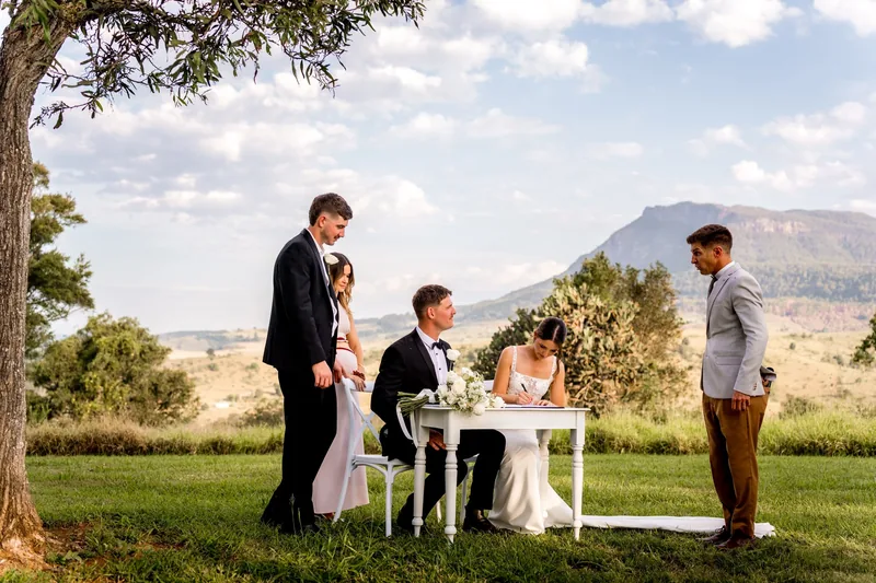 A couple signing at the white signing table on the lawn, the mountains behind