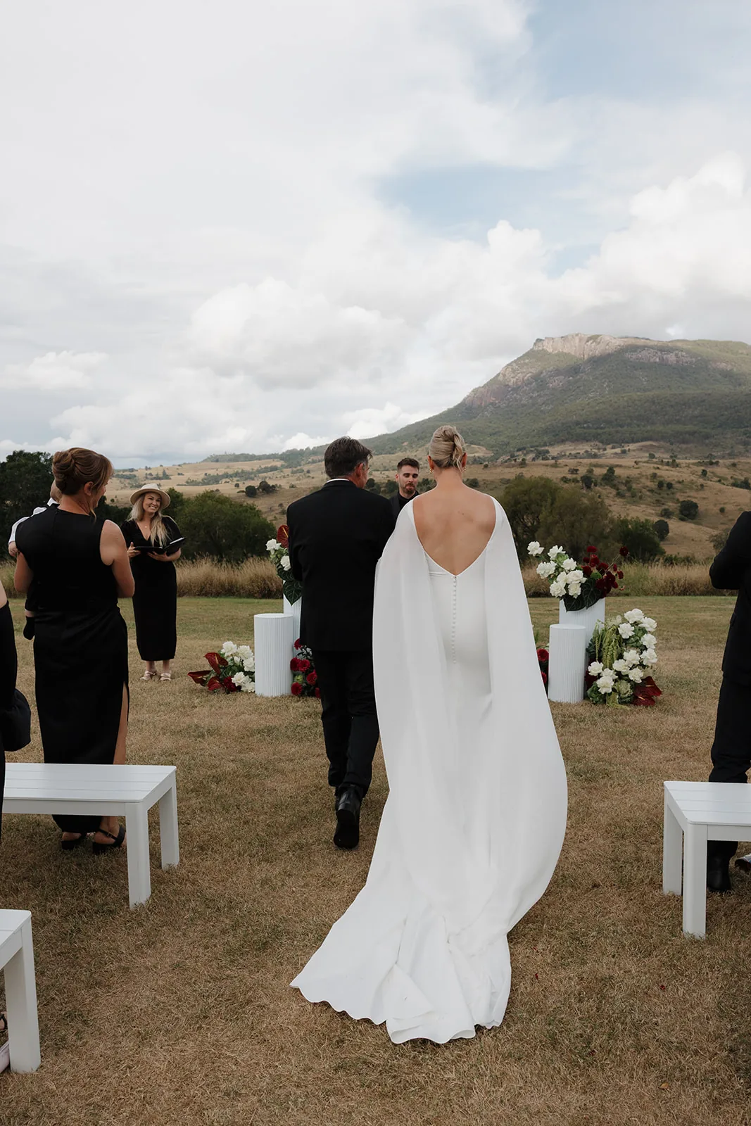 Jess walking down the aisle in a cape-back gown, mountain views behind