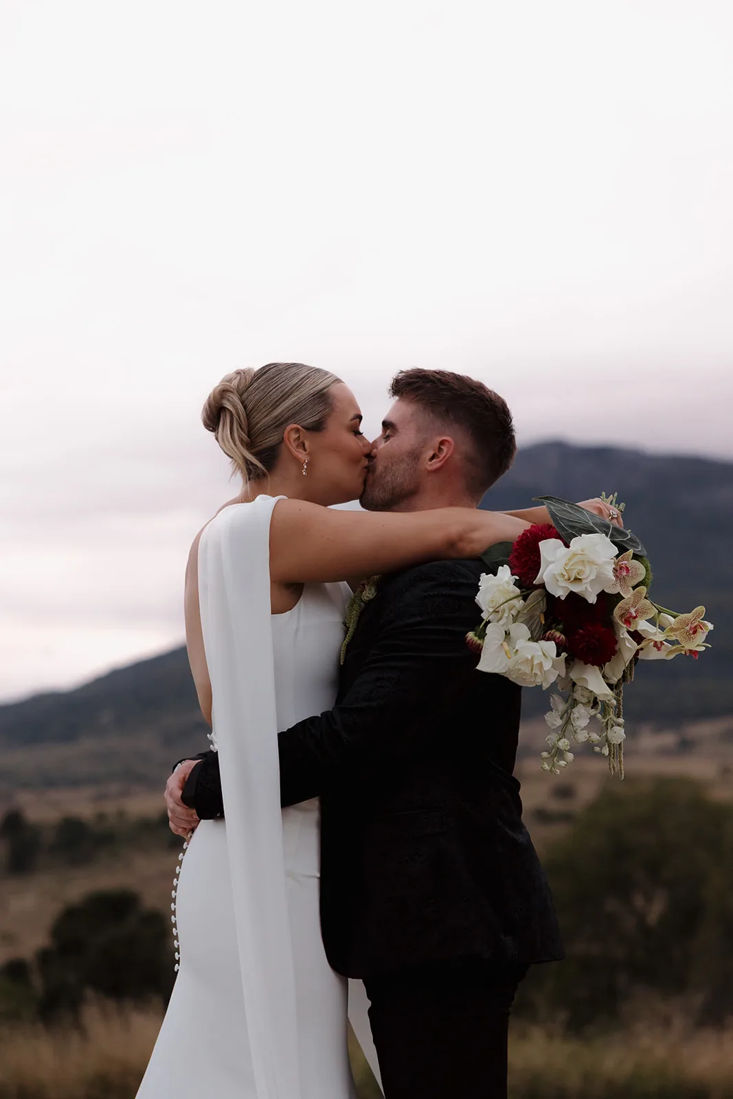 Jess and Cory sharing a kiss at dusk, holding her bouquet of white and burgundy flowers