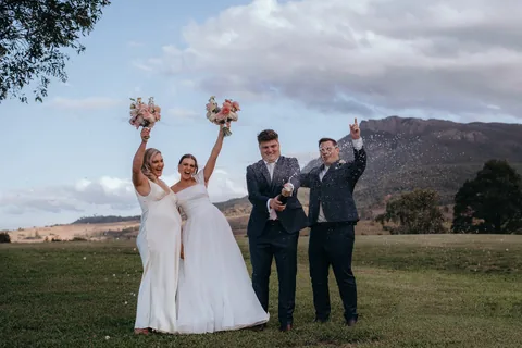 Two brides raising their bouquets as champagne sprays, blue sky and Mount Maroon behind