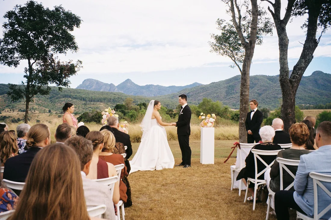Wide view of the ceremony in progress beneath gum trees, guests seated, mountains behind