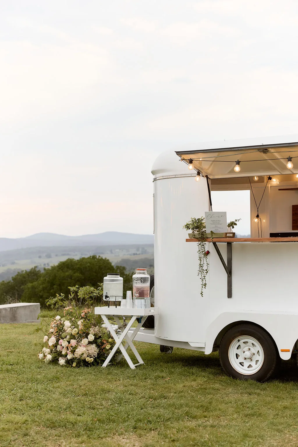 Mobile bar caravan lit up at dusk, styled with fresh florals and market lights