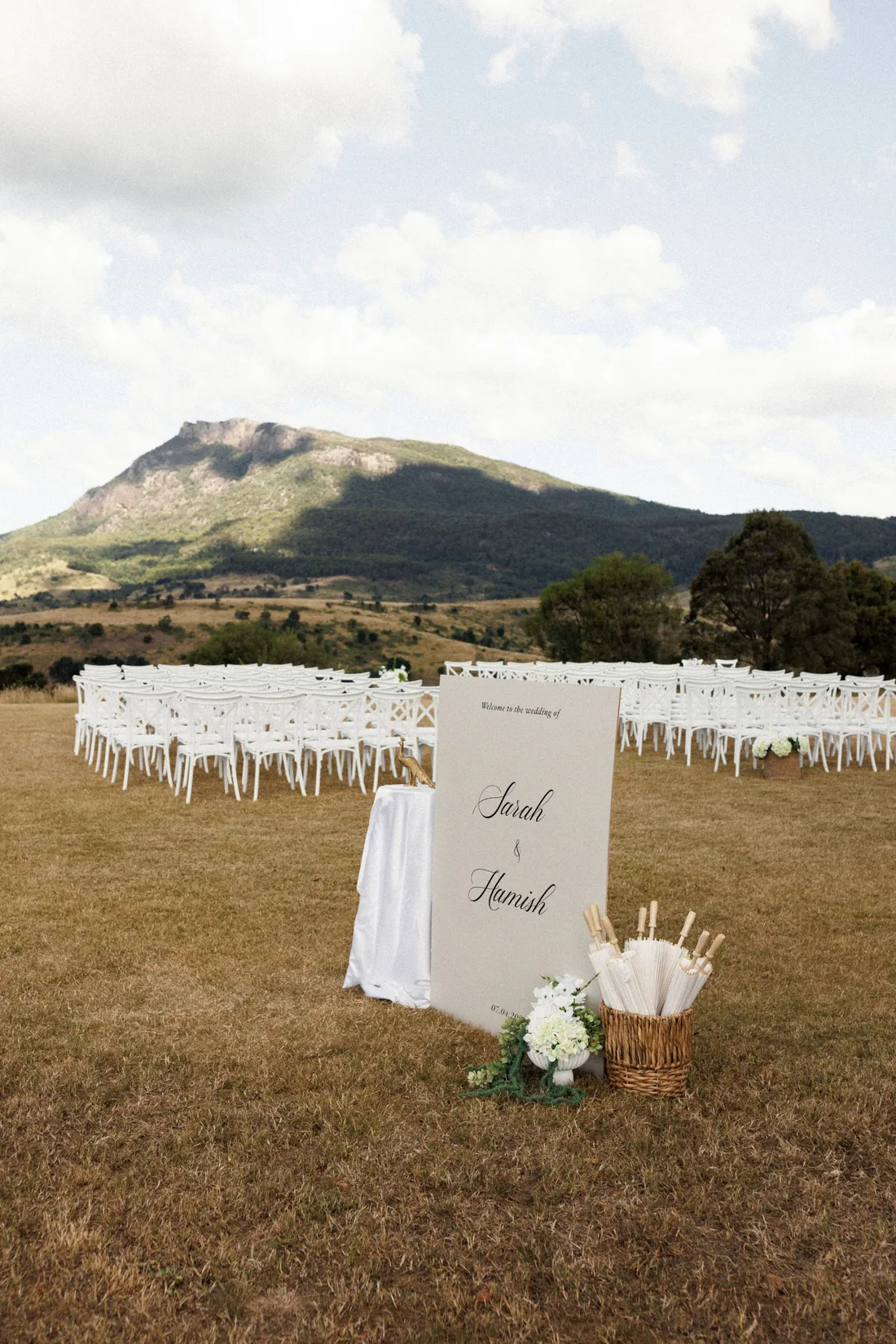 Sarah and Hamish wedding welcome sign on the lawn, rows of white chairs and mountain views behind