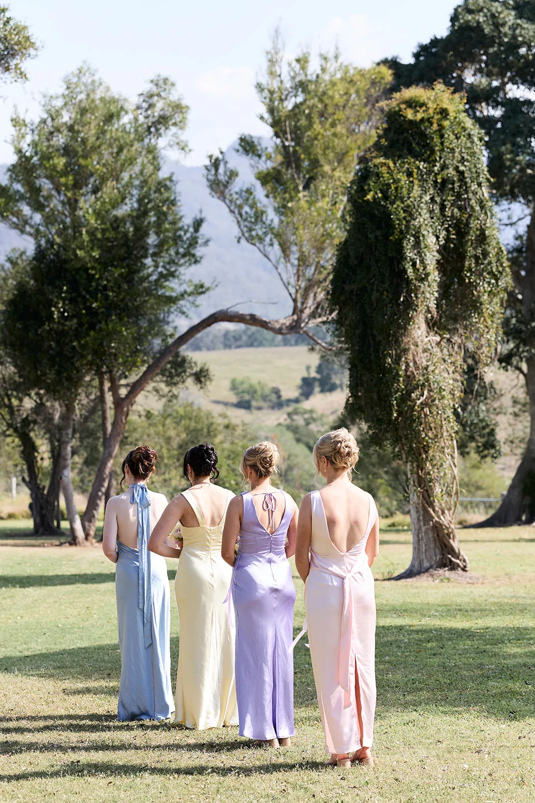 Bridesmaids in mismatched pastel gowns walking beneath a vine-draped tree after the ceremony