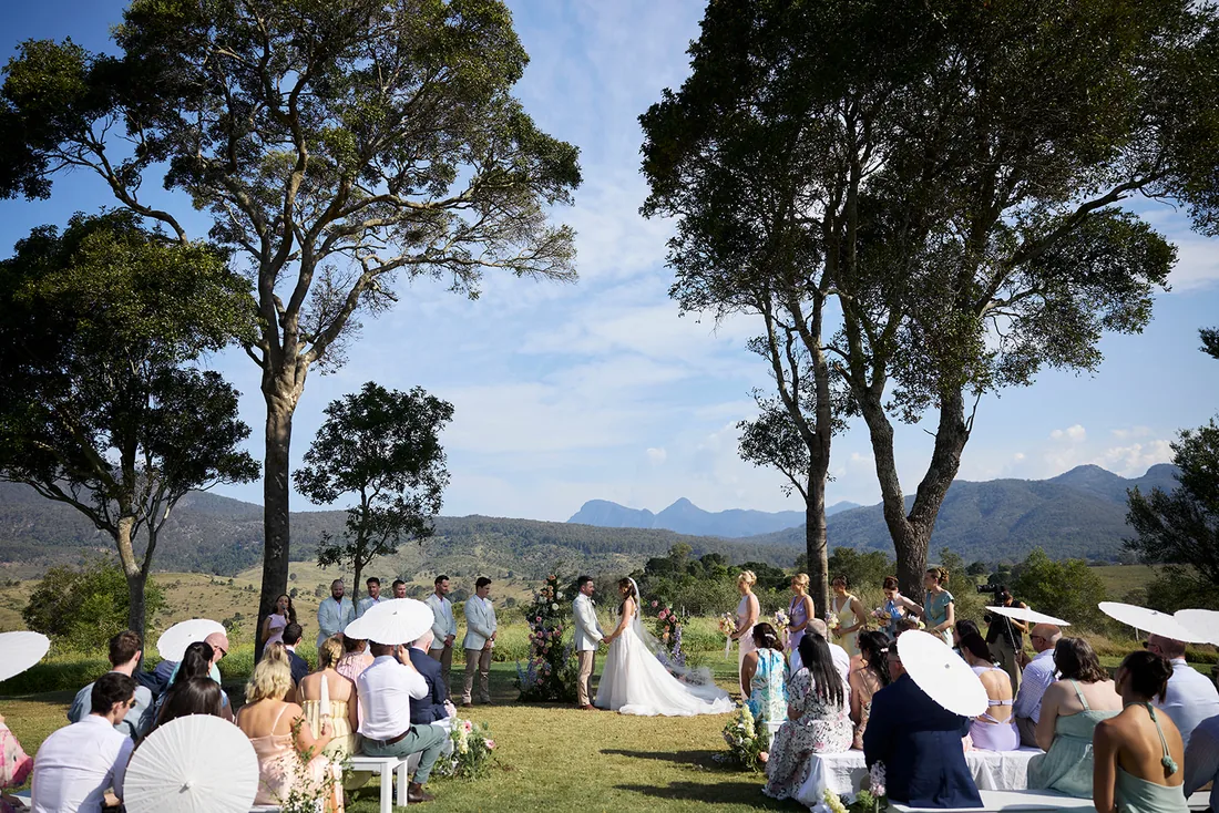 Wide view of Taylor and Jayden's ceremony beneath gum trees, guests seated under white parasols with mountains behind
