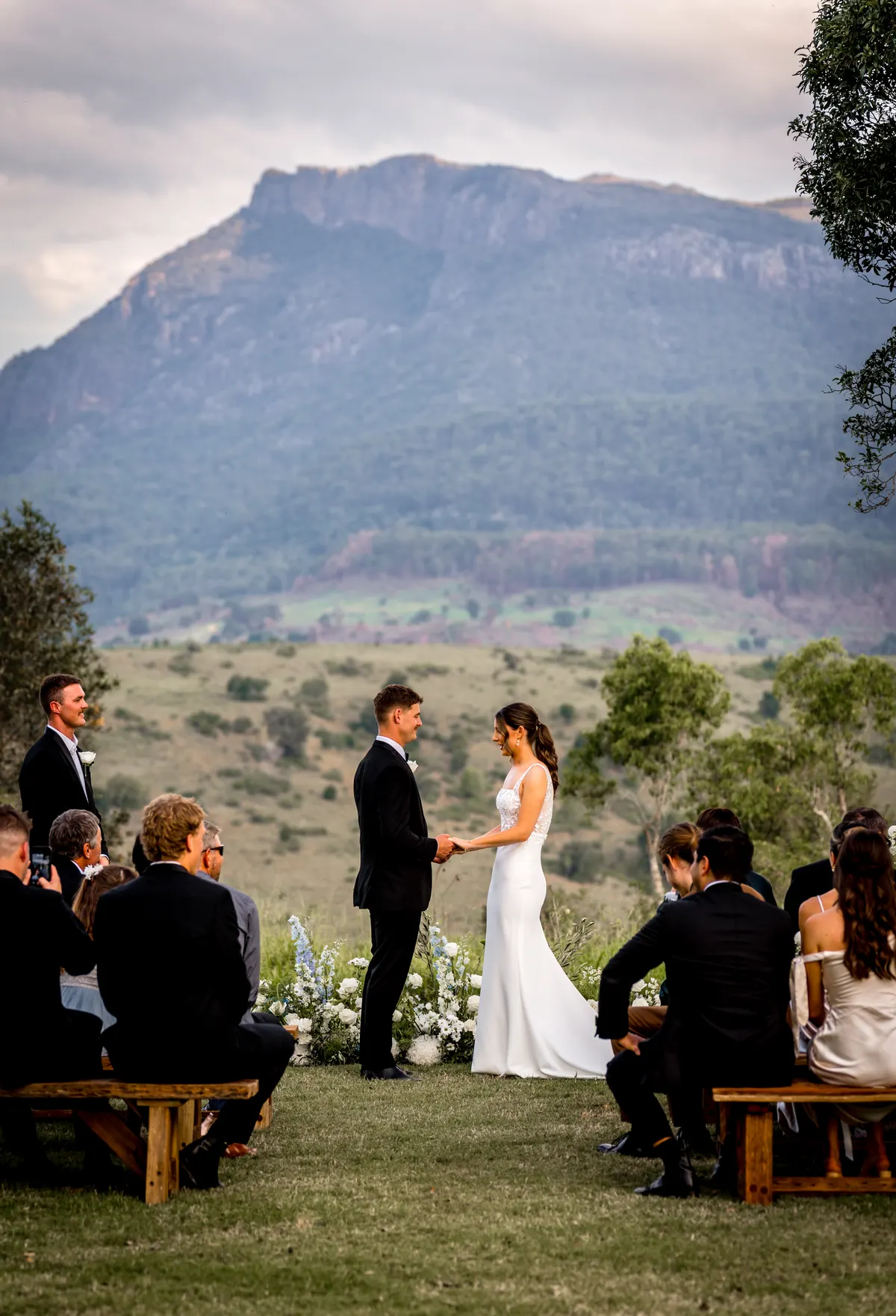 Bride and groom exchanging vows in front of guests seated on benches, with Mount Maroon rising behind them