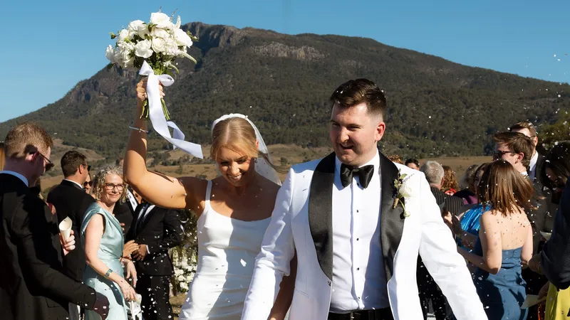 Siobhan and Ned walking back up the aisle through confetti, bouquet raised, Mount Maroon behind them